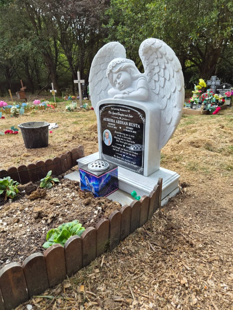 White marble angel-wing headstone with carved sleeping cherub, black inscribed plaque with ceramic portrait, flower vase and wooden picket grave surround at Hendon Cemetery and Crematorium, North London, memorial installation