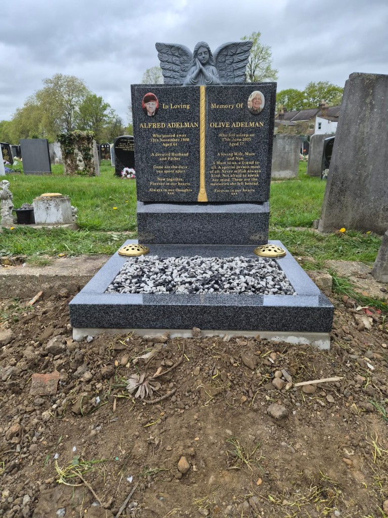 Polished dark speckled granite open book memorial with angel statue, ceramic portrait ovals, gold lettering and pebble-filled base with flower holders at Manor Park Cemetery, East London, memorial installation