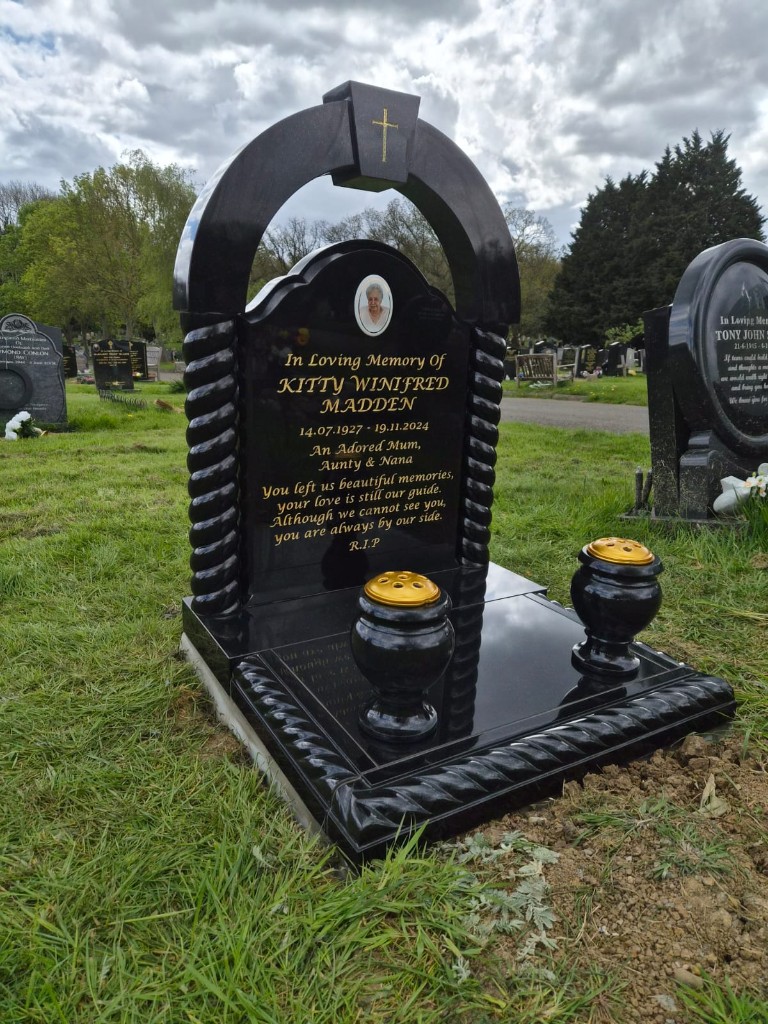 Polished black granite memorial with rope-carved arch pillars, gold cross and lettering, ceramic portrait oval and twin urn vases on tiered base at St Pancras and Islington Cemetery, North London, memorial installation