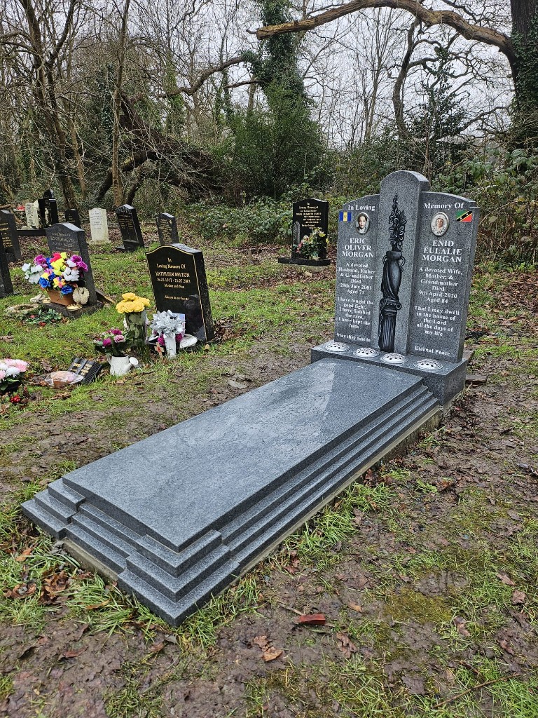 Polished dark grey speckled granite double memorial with twin portrait plaques, carved central vase motif, integrated flower holders and stepped full-length grave cover at St Pancras and Islington Cemetery, North London, memorial installation